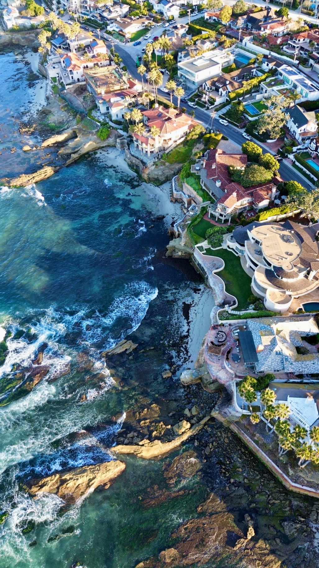 Aerial drone photograph looking straight down a rocky coastline with white surf breaking against the shore beside cliffside homes